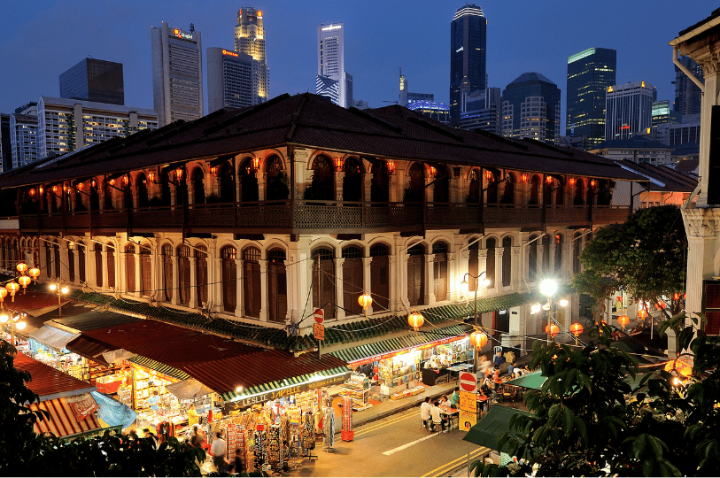 An elevated evening view of a historic multi-story shophouse in Chinatown, adorned with glowing red lanterns, with bustling street market stalls in the foreground and the modern Singapore skyline rising in the background.