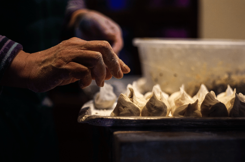 Dumpling- (5) A close-up, atmospheric shot of a pair of hands dusted with flour, carefully arranging a row of freshly pleated, raw dumplings on a metal tray in a dimly lit kitchen.