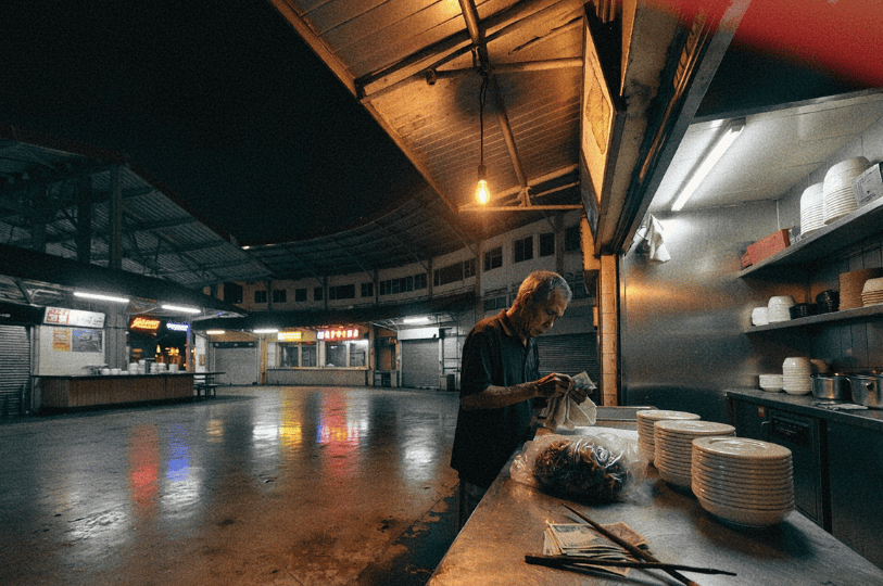 Final-Hours- (3) A cinematic, low-angle shot of an elderly vendor counting the day's earnings at a stainless steel counter. Stacks of clean white bowls sit ready for the next day. The background shows a vast, empty courtyard with colorful neon reflections on the wet ground under a dark night sky.