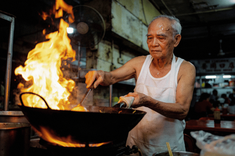 A portrait of an elderly hawker chef in a white singlet cooking with intense focus. He is tossing food in a large wok, creating a massive burst of flames (wok hei), capturing the sweat, labor, and soul of traditional Singapore street food culture.
