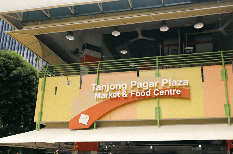 A low-angle shot of the Tanjong Pagar Plaza Market & Food Centre signage, featuring bright yellow and orange panels against the modern concrete architecture and lush green trees of the Singapore business district.