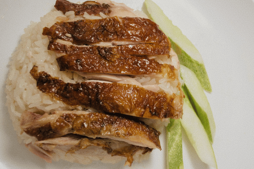 TanjongPagar- (3) A close-up of traditional Singaporean Hainanese chicken rice, showing succulent slices of chicken with glistening, charred bronze skin resting on a bed of fragrant oily rice, accompanied by fresh cucumber slices.