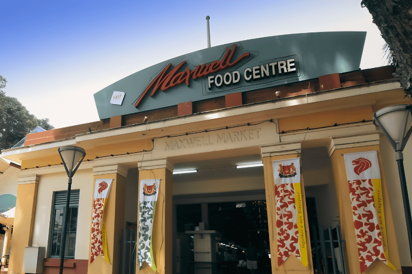TanjongPagar- (4) The historic facade of Maxwell Food Centre under a clear blue sky, featuring its iconic Art Deco-style entrance, mustard-yellow pillars, and celebratory red and white banners.