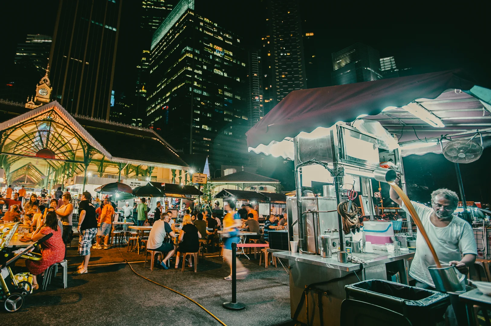 Vibrant night street food market with outdoor dining tables, food stalls, and illuminated city skyscrapers in the background.