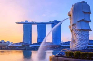 Wide-angle scenic shot of the iconic Merlion statue spouting water during golden hour, with the Marina Bay Sands silhouette in the background under a soft pastel morning sky.