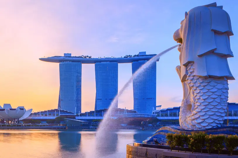 Wide-angle scenic shot of the iconic Merlion statue spouting water during golden hour, with the Marina Bay Sands silhouette in the background under a soft pastel morning sky.