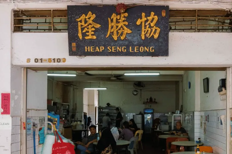 Breakfast-Spots- (3) Eye-level street photography shot of the rustic Heap Seng Leong kopitiam, capturing the moody, shadowed interior and the aged texture of its vintage gold-and-black signboard.
