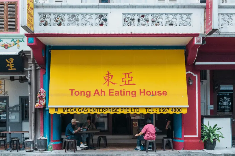 Breakfast-Spots- (6) A visually striking street-level photograph emphasizing the vibrant yellow awning and red Chinese typography of Tong Ah Eating House, casting a soft, diffused shade over alfresco diners.
