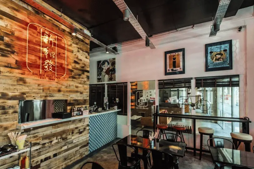 Breakfast-Spots- (8) Moody interior shot of Sin Hoe Huat Cafe, focusing on the warm, ambient glow of a red neon sign mounted on a textured wooden pallet wall, with glossy black tables catching the light.