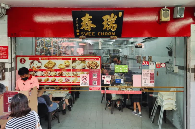Breakfast-Spots- (9) Brightly lit, straight-on capture of the Swee Choon restaurant entrance, showcasing its striking red and gold overhead sign and glass doors heavily decorated with colorful dim sum menus.