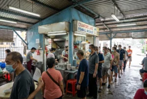 A wide shot capturing a long line of customers waiting patiently in front of the Crawford Lane Fishball Noodles stall at a bustling Singapore hawker centre.