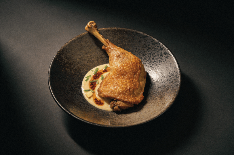 Imperial-Diversity- (3) A high-contrast, dramatic studio shot of an elevated fine-dining dish, featuring a perfectly crisped duck leg resting on a creamy sauce dotted with chili oil and fresh chives in a dark ceramic bowl.