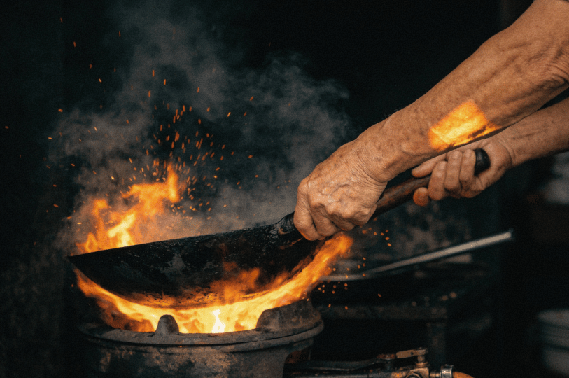 Imperial-Diversity- (4) A dynamic, close-up shot of an older chef's arms as they vigorously toss a seasoned black wok over a roaring open flame, capturing the glowing fire and flying sparks of traditional wok cooking.