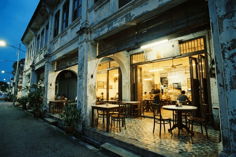 An exterior wide shot of a traditional Singaporean shophouse restaurant at dusk. The blue hour sky contrasts with the warm, inviting yellow glow spilling from the open folding doors onto the patterned tile sidewalk and outdoor marble-top dining sets.