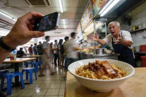 A first-person perspective shot of a diner holding a smartphone to photograph a bowl of noodles in the foreground. In the background, an elderly hawker vendor gestures expressively with open arms toward the camera, while a blurred line of customers forms behind him.