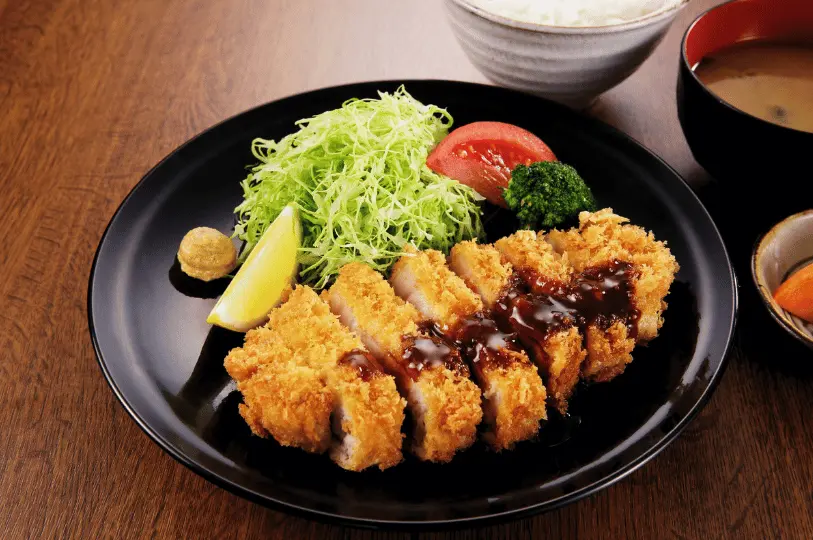 A classic Japanese tonkatsu set served on a sleek black plate, featuring sliced breaded pork drizzled with dark sauce, a mountain of finely shredded cabbage, a lemon wedge, and bowls of rice and miso soup in the background.