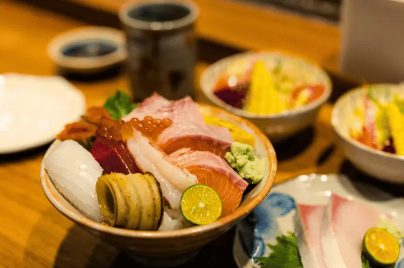 A close-up view of a colorful Japanese chirashi bowl topped with assorted fresh sashimi, salmon roe, squid, and a slice of citrus, served on a wooden counter.
