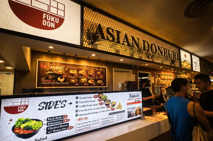 The brightly lit storefront of the Fuku Don Asian Donburi food stall, showing customers gathered at the counter under glowing digital menu boards.
