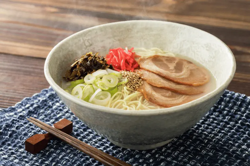 A steaming bowl of tonkotsu ramen served in a textured ceramic bowl on a blue woven placemat, garnished with chashu pork, green onions, sesame seeds, wood ear mushrooms, and bright red pickled ginger.