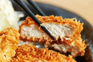 Close-up of black chopsticks lifting a thick slice of crispy, golden-brown tonkatsu, revealing the juicy, tender white pork inside.