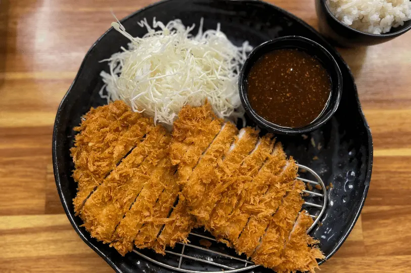 A generous portion of freshly fried, sliced tonkatsu resting on a semi-circular wire cooling rack to preserve its crispiness, served alongside a pile of shredded cabbage, a bowl of sesame-flecked dipping sauce, and steamed white rice.