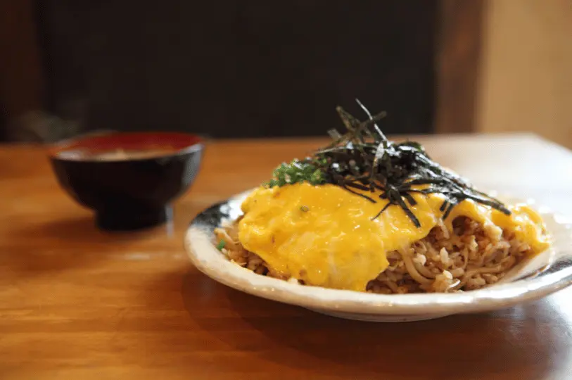 A side-angle, shallow depth-of-field photograph of a comforting plate of omurice topped with a bright yellow omelet and shredded nori, resting on a wooden table with a softly blurred bowl of miso soup in the background.