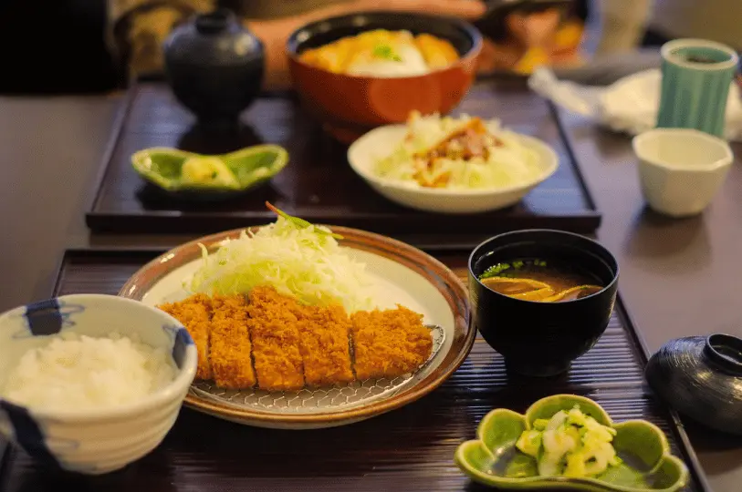 A classic Japanese tonkatsu set meal served on a dark wooden tray, featuring a crispy breaded pork cutlet, shredded cabbage, white rice, pickles, and a bowl of miso soup.