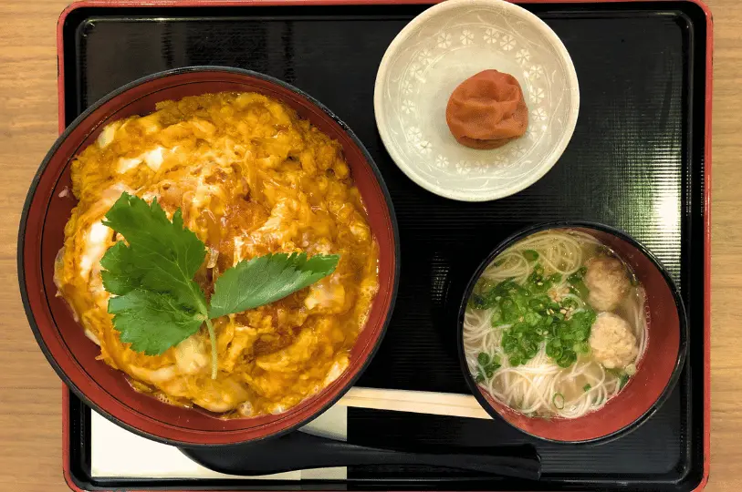 A top-down view of a classic Japanese set meal on a black tray, featuring a vibrant bowl of katsudon garnished with fresh green mitsuba, a side of clear somen noodle soup with meatballs, and a small dish holding a single pickled plum.