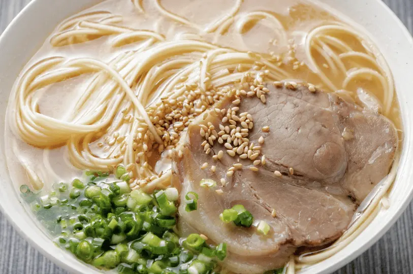 An overhead close-up of a bowl of tonkotsu ramen showing straight noodles resting in a milky, fatty broth, topped with a large slice of chashu pork, toasted sesame seeds, and chopped green onions.