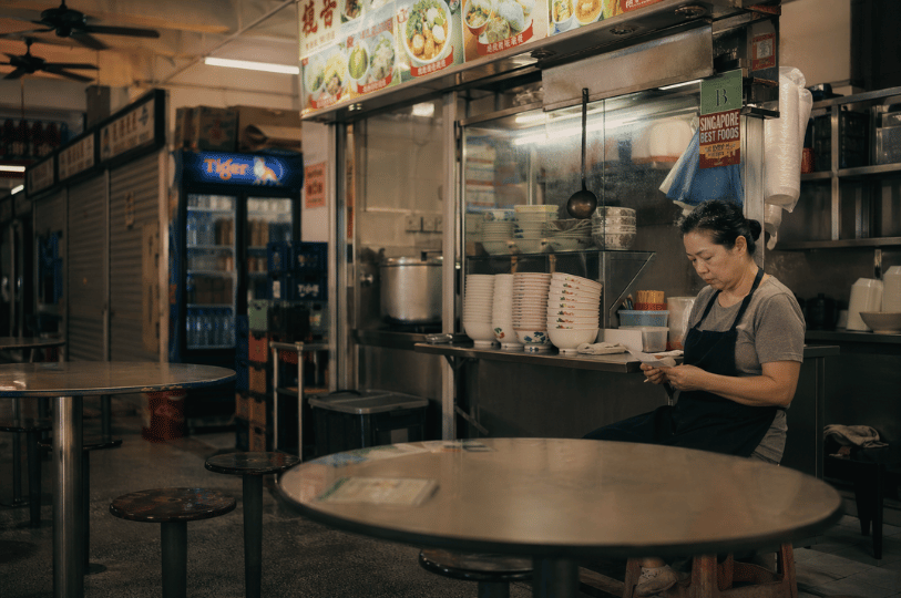 An older female hawker wearing a dark apron sits alone at a round metal table in a dimly lit, quiet food centre, looking down at a small piece of paper in her hands. Behind her is her stall, featuring neat stacks of bowls on a stainless steel counter, an illuminated Tiger beer refrigerator in the background, and a menu board displaying pictures of noodle dishes.