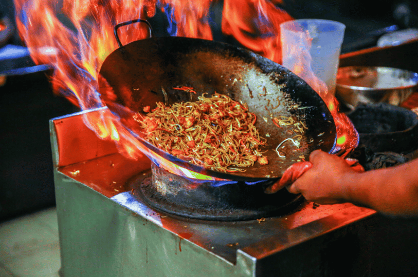 Close-up of the hawker's hands steadily tossing thick noodles and vegetables in a heavy wok. Fierce orange and blue flames from a commercial gas burner roar upward, licking the edges of the dark metal. The background is blurred into a soft wash of light, keeping the focus entirely on his hands and the intense heat of his quiet, practiced labor.
