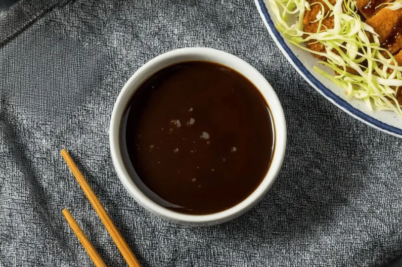 A top-down view of a small white bowl filled with thick, glossy, dark brown tonkatsu sauce, resting next to wooden chopsticks on a textured grey cloth.