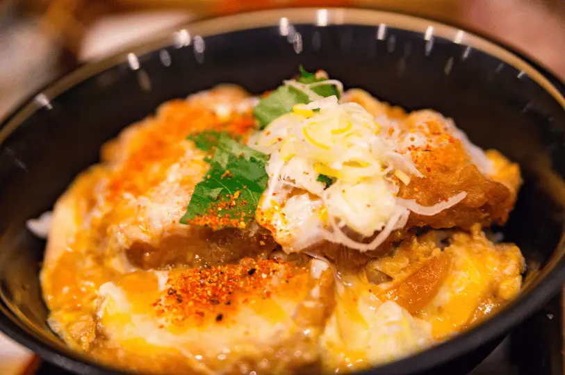 A close-up food photography shot of a hearty katsudon bowl with a shallow depth of field, highlighting the savory breaded pork cutlet bound by soft egg, garnished with chopped scallions, and dusted with shichimi togarashi under warm, appetizing lighting.