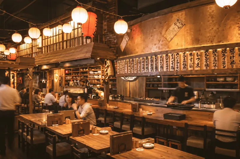 A warm, traditional-style izakaya interior filled with wooden tables, glowing paper lanterns, and hanging wooden menu boards, with customers chatting over dinner and drinks.
