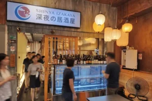 Diners walking through the brightly lit entrance of Shinya Izakaya in Singapore, featuring hanging paper lanterns and a glass display case lined with sake bottles.