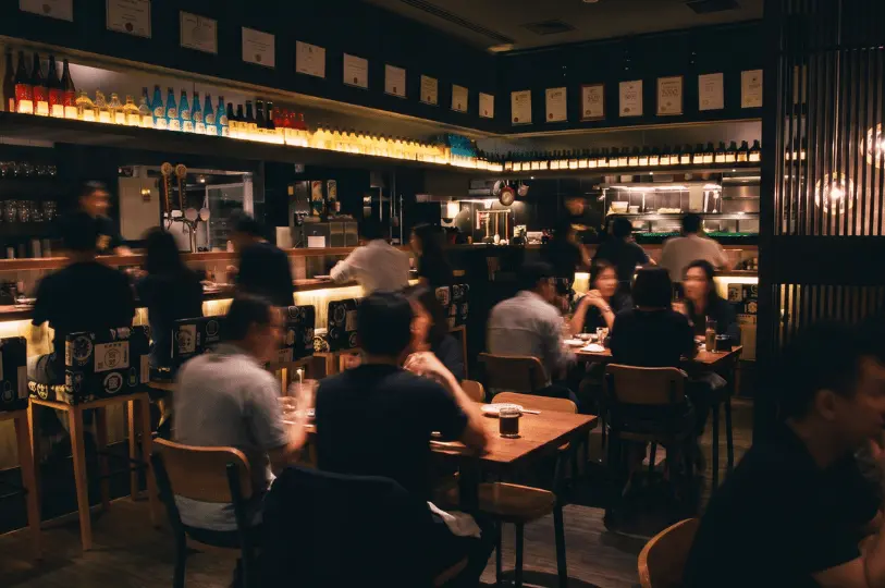 The lively, dimly lit interior of a bustling izakaya filled with patrons dining at wooden tables and a bar counter, framed by a warmly glowing display of backlit sake bottles lining the upper shelves.