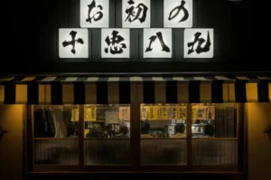 A warmly lit storefront of a traditional izakaya at night, featuring illuminated Japanese calligraphy signs, a striped awning, and the silhouettes of patrons dining at the counter visible through the front windows.