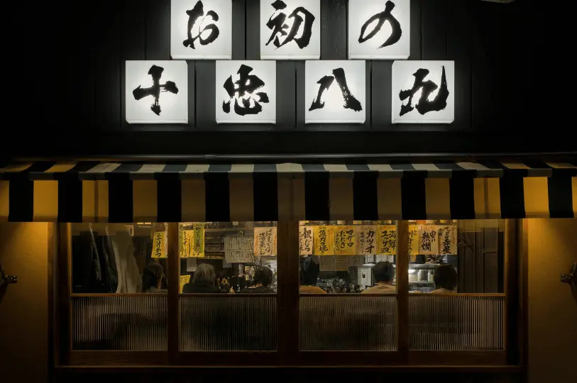 A warmly lit storefront of a traditional izakaya at night, featuring illuminated Japanese calligraphy signs, a striped awning, and the silhouettes of patrons dining at the counter visible through the front windows.