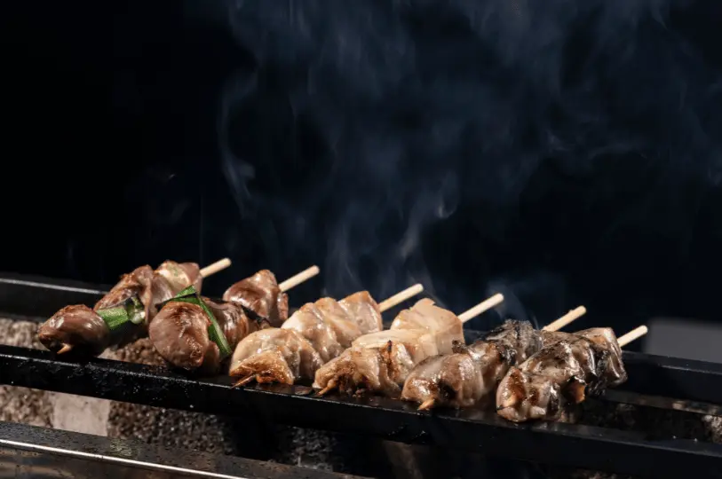 Several yakitori skewers cooking on a metal grill grate, with thick, dramatic plumes of white smoke rising from the meat against a stark black background.