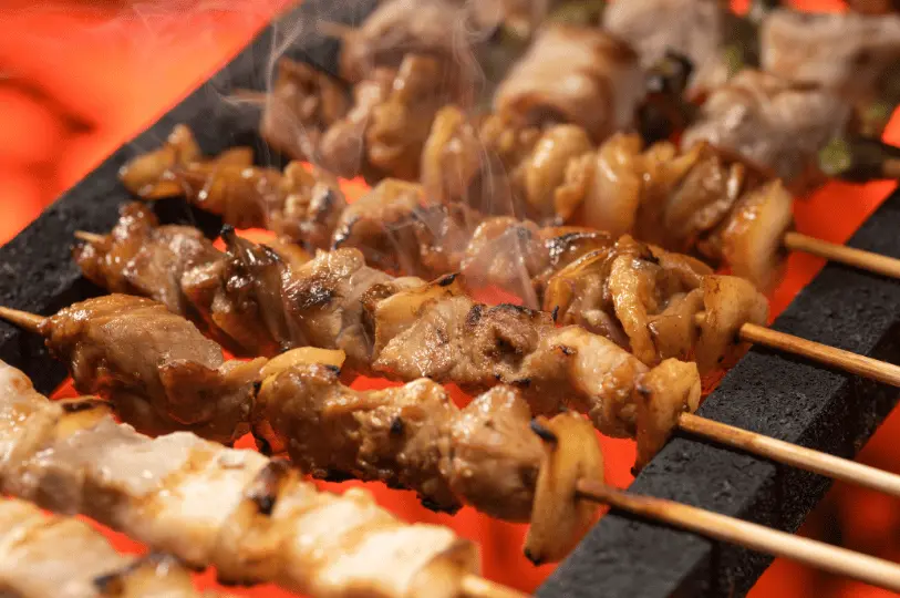 A close-up of multiple chicken yakitori skewers sizzling on a grill directly over glowing, intensely red-hot charcoal, with light smoke rising from the glistening meat.