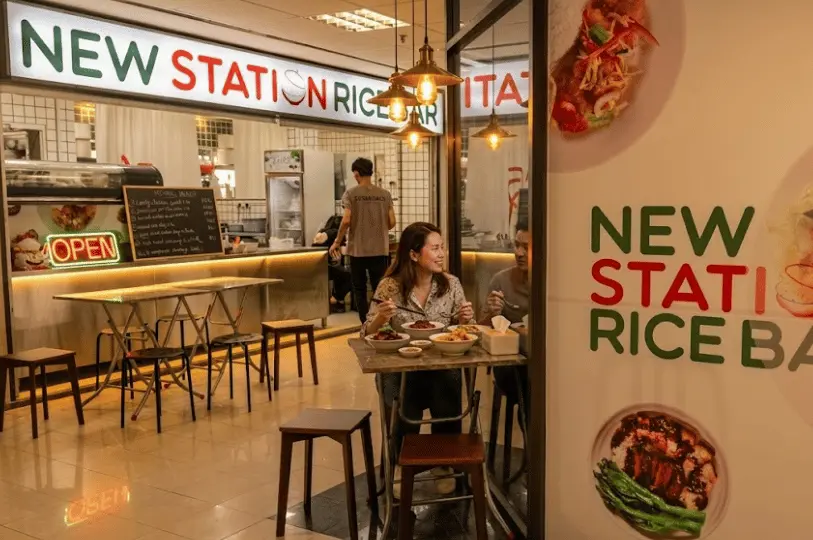 Two patrons smiling and enjoying a shared meal together at a small table just outside a lively eatery, illuminated by dramatic, warm pendant lighting.