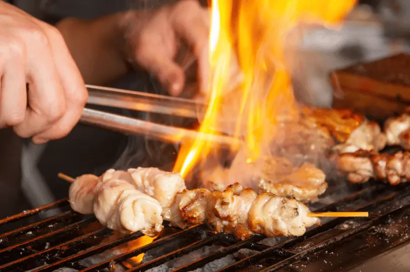 A chef's hands using metal tongs to turn yakitori skewers on a charcoal grill, as bright orange flames aggressively flare up to sear the meat.