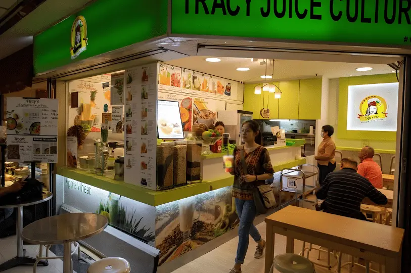 A woman walks past a vibrant, green-themed juice and beverage stall holding a freshly made drink, with other diners casually seated nearby in a warmly lit setting.
