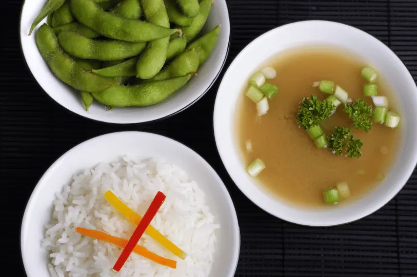 A top-down view of classic izakaya side dishes served in white bowls against a dark bamboo mat, featuring steamed edamame, white rice with a bell pepper garnish, and clear soup with scallions.