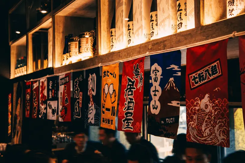 The lively interior of an izakaya showing a row of colorful hanging banners with traditional Japanese designs and calligraphy, positioned right below a wooden shelf lined with illuminated sake bottles.