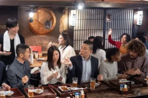 A lively scene inside a warm, wood-paneled izakaya where a chef with a towel around his neck happily interacts with a diverse group of patrons seated at the bar.