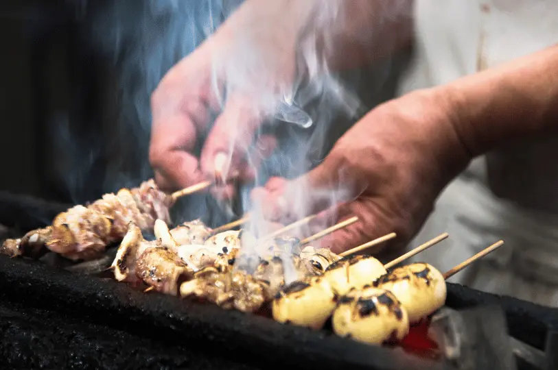 A detailed, smoky close-up of meat and vegetable skewers sizzling on a hot charcoal grill, capturing the dynamic movement of thick white smoke rising between the chef's hands as they turn the food.