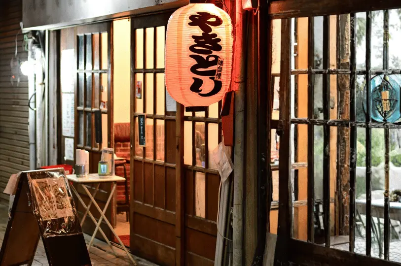 The inviting exterior of a local izakaya featuring traditional wooden sliding glass doors, a sidewalk menu board, and a brightly lit red paper lantern hanging welcomingly at the entrance.