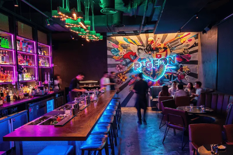 A trendy, modern izakaya interior with vibrant neon lighting, a long wooden bar counter, and a striking, colorful wall mural featuring a glowing neon 