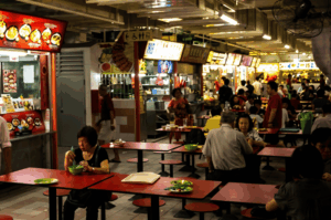 A bustling, crowded hawker centre capturing the relentless, everyday energy of the food market. In the foreground, patrons sit at scattered red tables enjoying their meals, while the background reveals a long, brightly illuminated row of food stalls where busy vendors work continuously to serve the steady flow of customers.